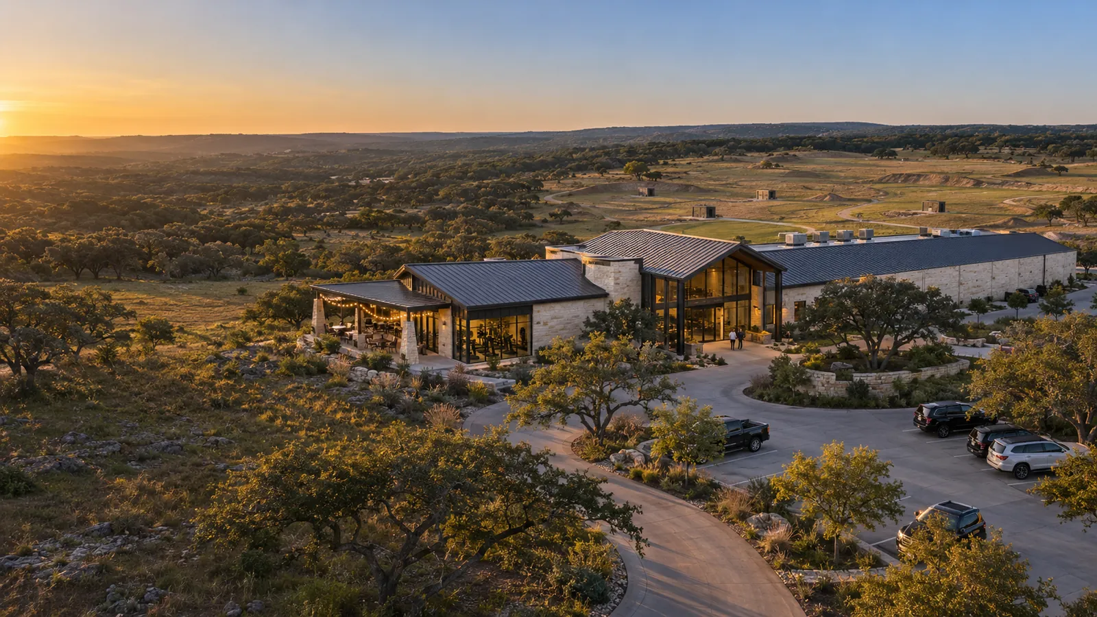 A stone and steel clubhouse at sunset in the Texas hill country, with a long range wing, a dining patio and a clays field beyond